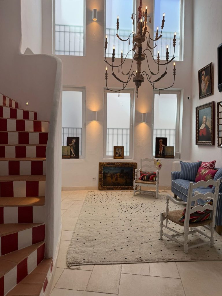 Salón de doble altura con ventanales, lámpara de araña de hierro y biombo de cuadros rojo y blanco — proyecto de interiorismo en Marbella