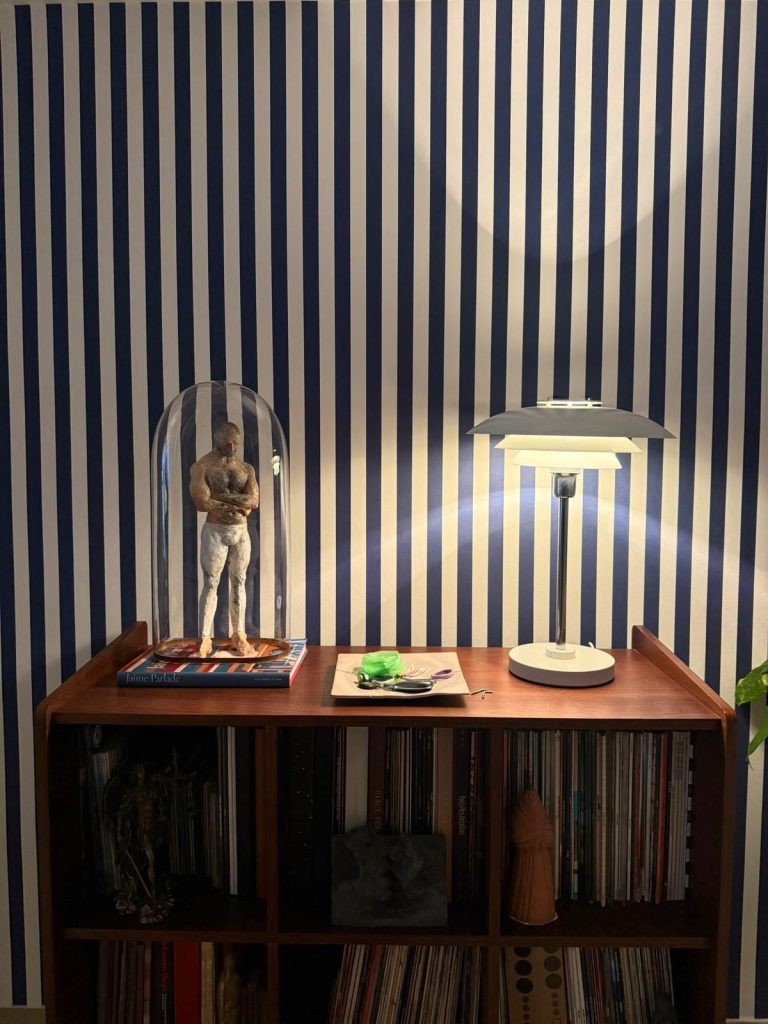 Mesilla de noche con lámpara de sobremesa y figura bajo campana de cristal frente a papel pintado de rayas azules